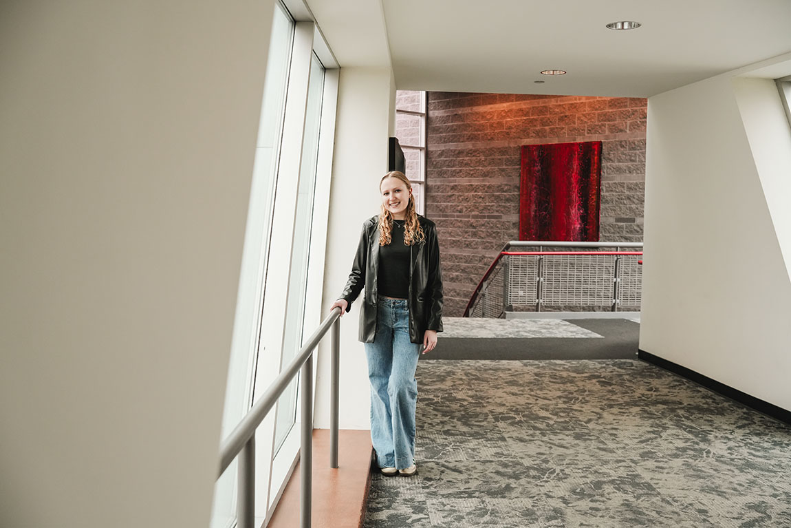 A college student wearing a black shirt and jacket smiles inside the corridor of an arts building